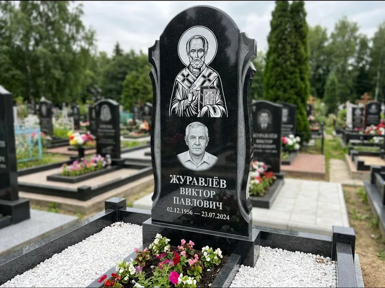 Памятник с гравировкой иконы: стела с образом в полный рост на обороте — гравировка образа Матроны Московской, низкие кусты спиреи в цвету, работа «Лебедева» в Якутске